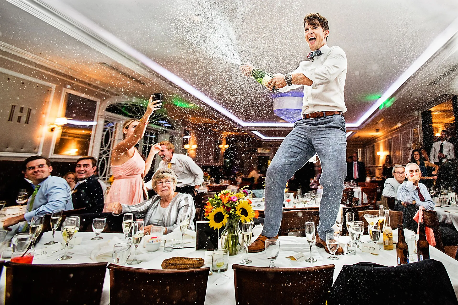 Wedding reception photo of a man standing on a table spraying champagne over the crowd, hero image from Unbridled photo book by Todd Laffler.